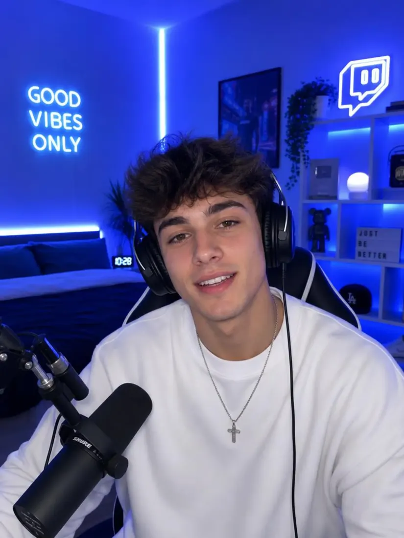 Young streamer in headphones smiling at a microphone in a room with blue lights and a Good Vibes Only sign