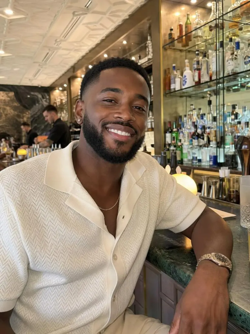 Smiling man in a cream textured knit shirt sitting at a bar with shelves of bottles