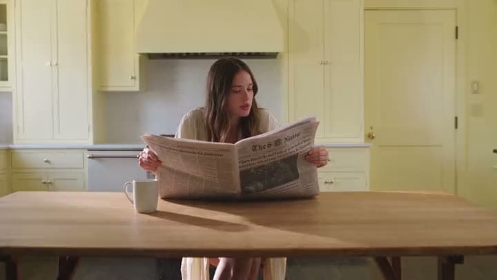 A woman reading a newspaper at a wooden table in a pale yellow kitchen.