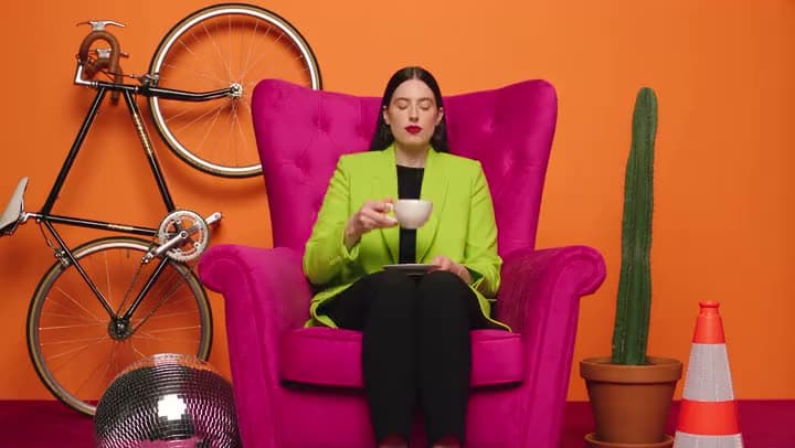 A woman sipping from a cup in a pink armchair, with a bike, a cactus and a traffic cone around her.