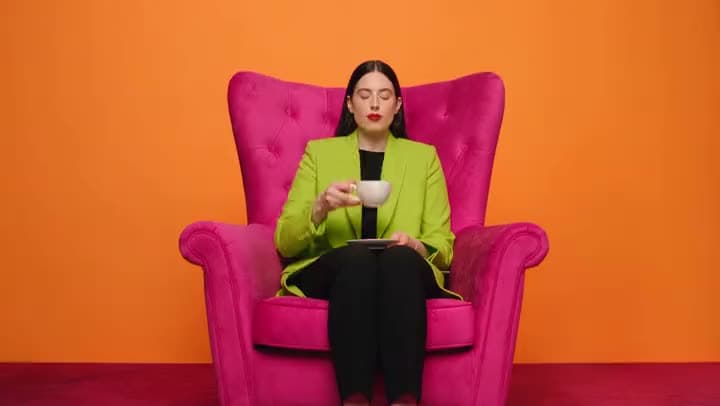 An AI video of a woman in a lime green blazer sipping from a teacup in a pink armchair against a bright orange wall.