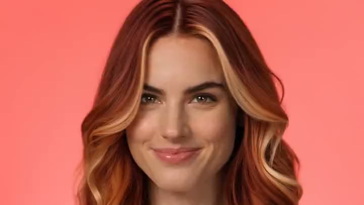 An AI video of a smiling woman with long wavy auburn hair and blonde highlights posed against a coral pink background.