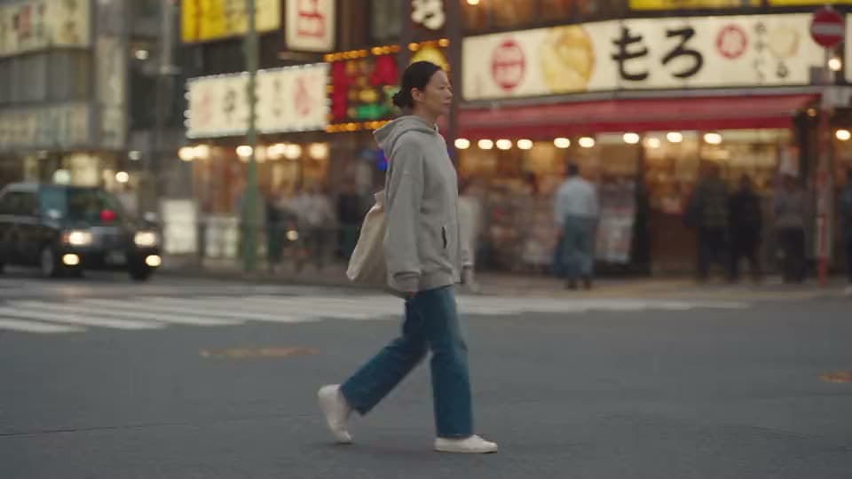 A woman in a grey hoodie and jeans crossing a street lined with Japanese shop signs.