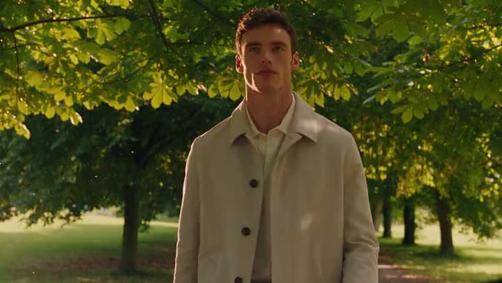 A man in a cream trench coat walking under green summer trees in a park.