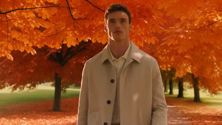 An AI video of a man in a beige overcoat walking through a park under trees with bright orange autumn leaves.