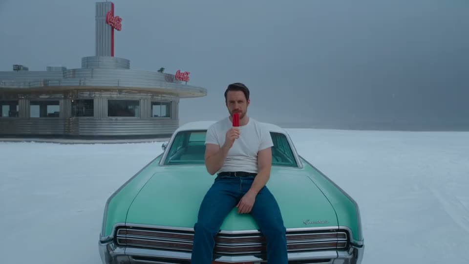 An AI video of a man in a white T-shirt eating a red popsicle on the hood of a teal car in the snow outside a Coca-Cola diner.