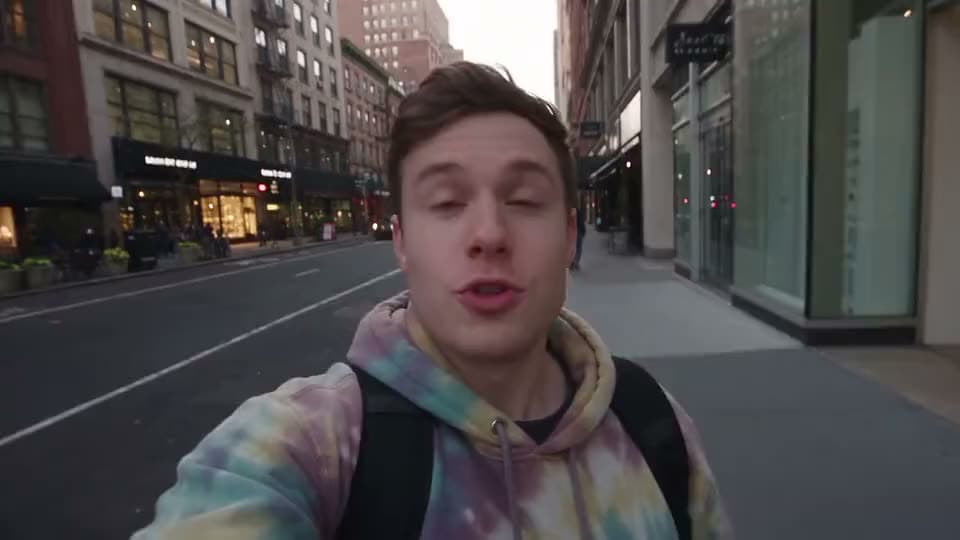 An AI video of a young man in a tie-dye hoodie filming himself as he walks down an empty city street at dusk.