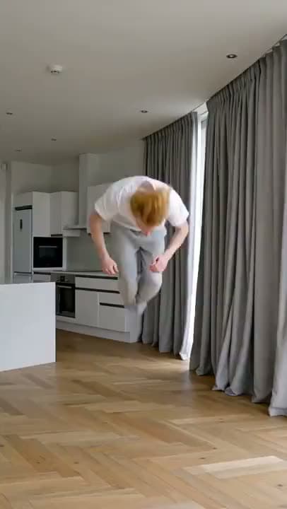 A vertical AI video of a red-haired man in a white T-shirt mid-backflip in a bright modern kitchen and living room.