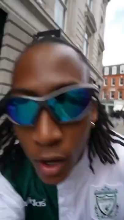 A vertical AI video of a man in wraparound blue sunglasses leaning into a selfie camera on a city street.