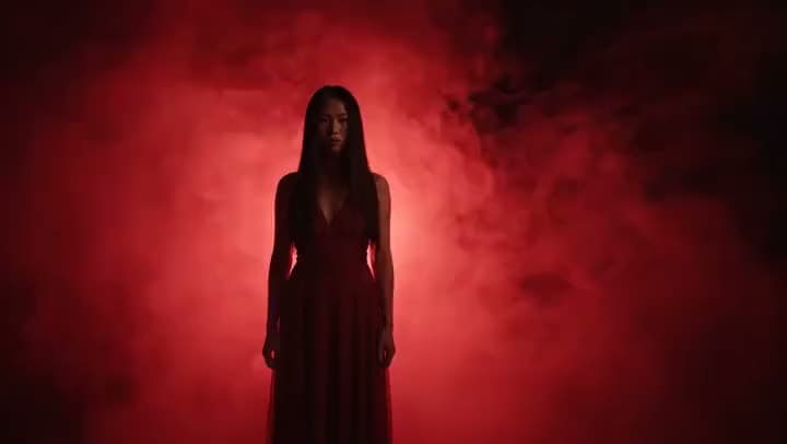 An AI video of a woman with long black hair in a dark red gown standing still in a cloud of glowing red smoke.