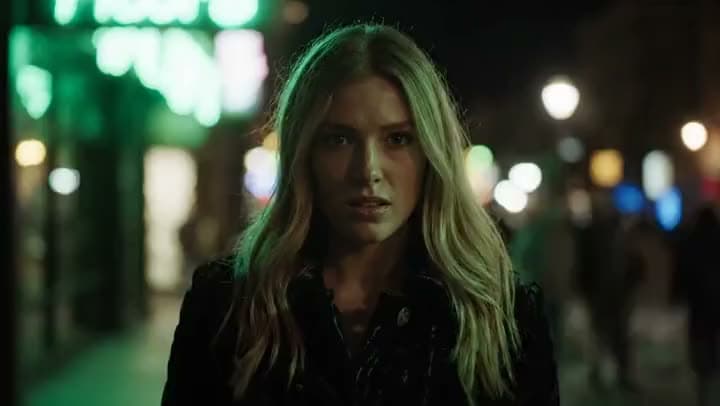 An AI video of a blonde woman in a dark jacket speaking to the camera on a city street at night, with neon lights blurred behind her.
