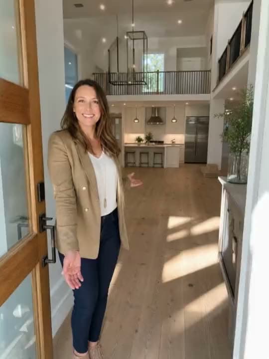 A vertical AI video of a real estate agent in a tan blazer welcoming viewers through the front door of a bright open home.