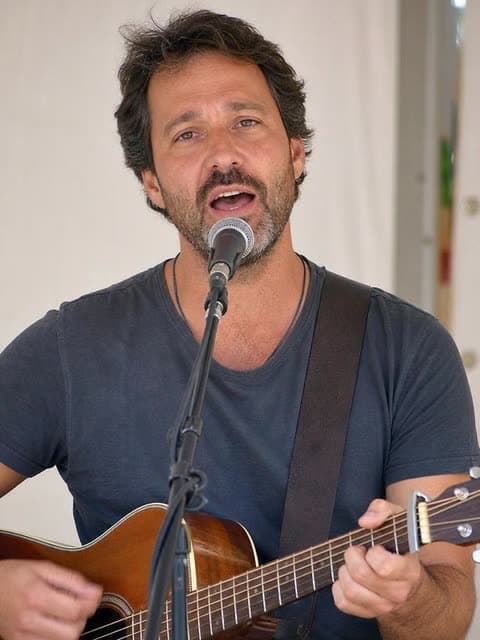 A portrait photo of a bearded man in a gray T-shirt singing into a microphone while playing an acoustic guitar.