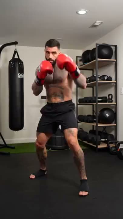 A vertical video of a tattooed boxer in red gloves and black shorts shadowboxing in a home gym beside a heavy bag.
