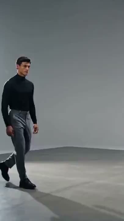 A vertical video of a man in a black turtleneck and gray trousers walking across a plain gray studio floor.