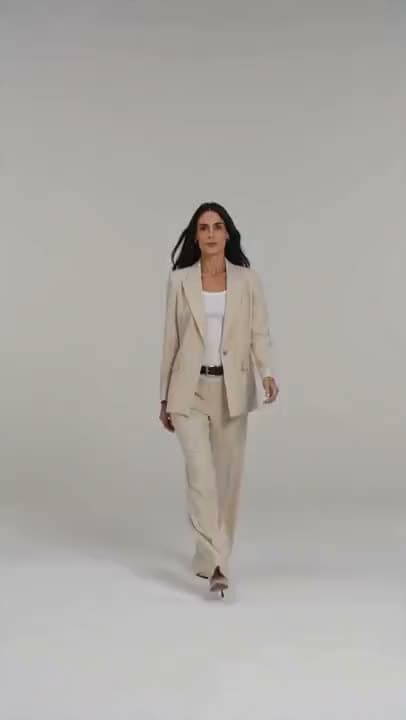 A vertical video of a dark-haired woman in a cream pantsuit walking toward the camera against a plain gray backdrop.