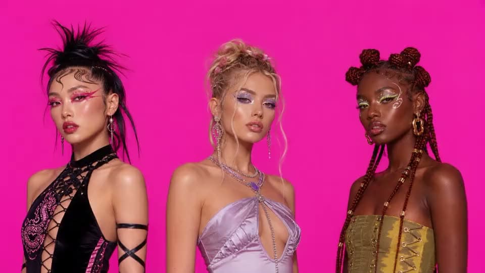An AI fashion video of three models in bold eye makeup and satin and corset tops posing against a hot pink backdrop.