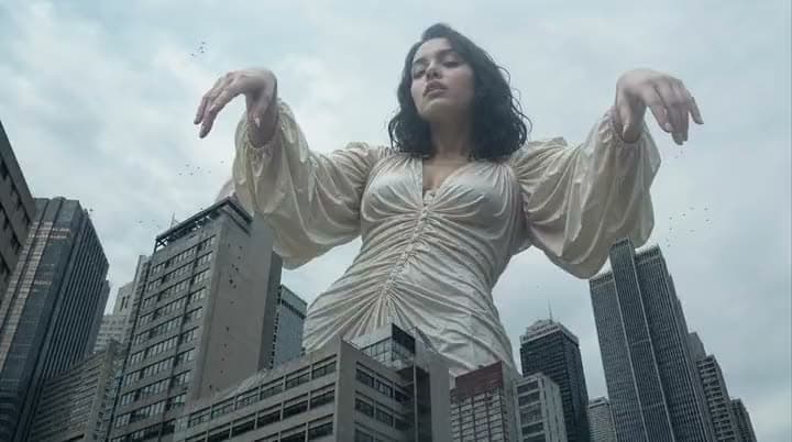An AI video of a giant woman in a cream satin dress towering over city skyscrapers with her arms raised.
