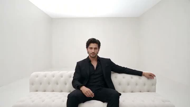 An AI video of a man in a black suit sitting on a white tufted sofa in the middle of an empty all-white room.