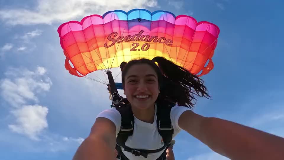 An AI video of a smiling woman filming a selfie mid-flight under a pink and orange parachute printed with "Seedance 2.0".