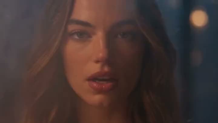 An AI music video close-up of a woman with long brown hair singing in warm, hazy light with soft bokeh behind her.