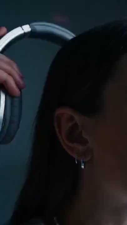 A vertical AI video close-up of a woman with a hoop earring as a hand lowers a pair of silver over-ear headphones onto her head.