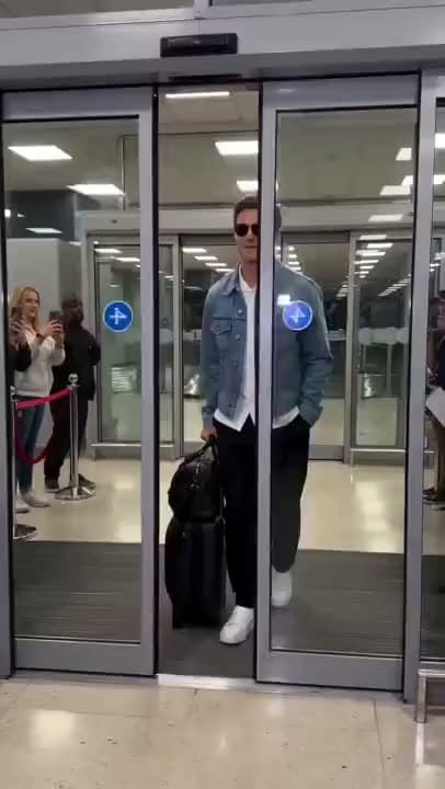 A vertical AI video of a man in sunglasses and a denim jacket walking out of airport doors with a bag as fans film him.