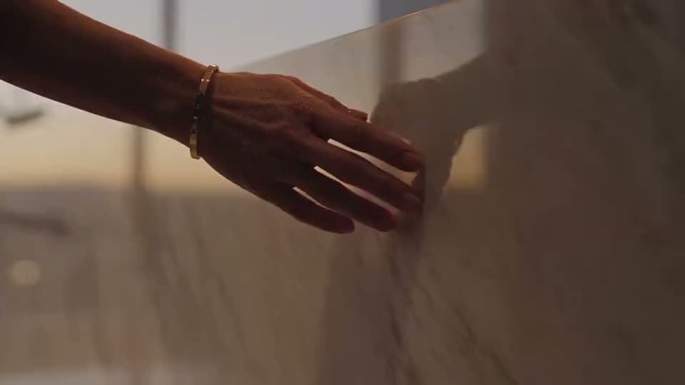 An AI video close-up of a woman's hand with a gold bracelet brushing along a marble wall in warm evening light.