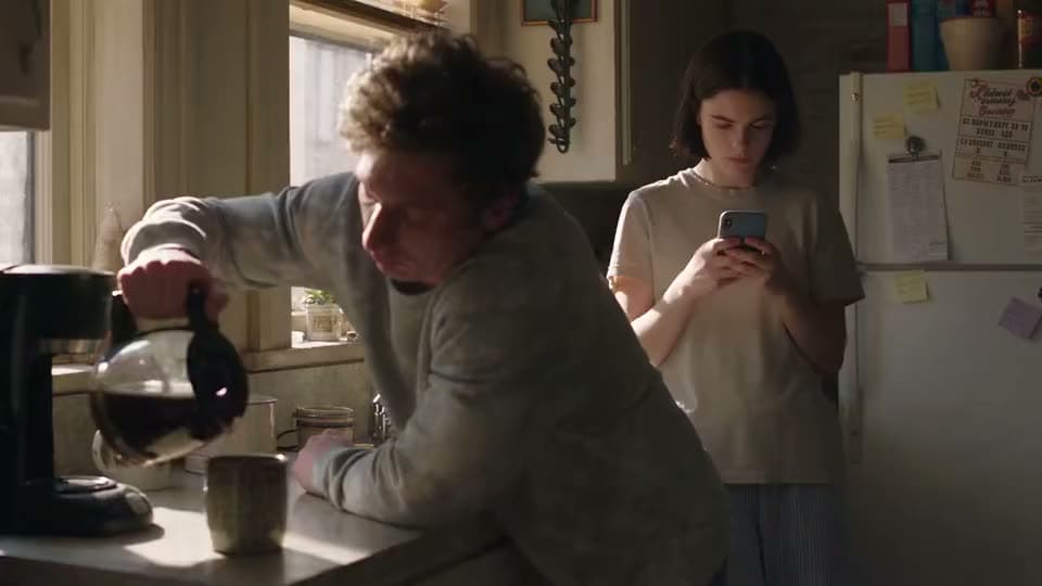 An AI video of a man pouring coffee in a sunlit kitchen while a teenage girl behind him looks down at her phone.
