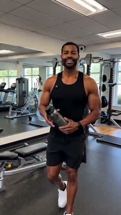 A vertical AI video of a smiling man in a black tank top holding a water bottle as he walks through a gym.