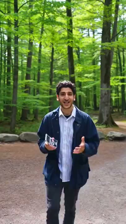 A vertical AI video of a smiling man in a navy jacket holding a small gadget as he talks to camera on a forest path.