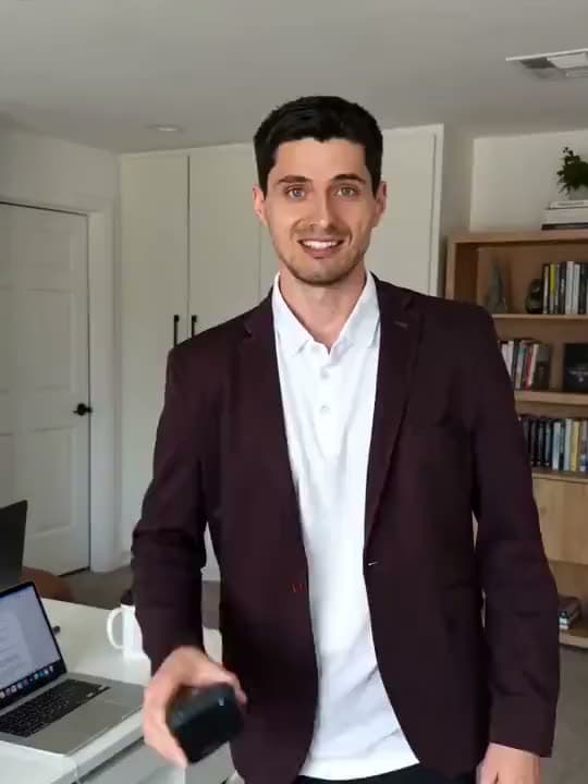 A vertical AI video of a smiling man in a burgundy blazer holding out a small black gadget in a home office.