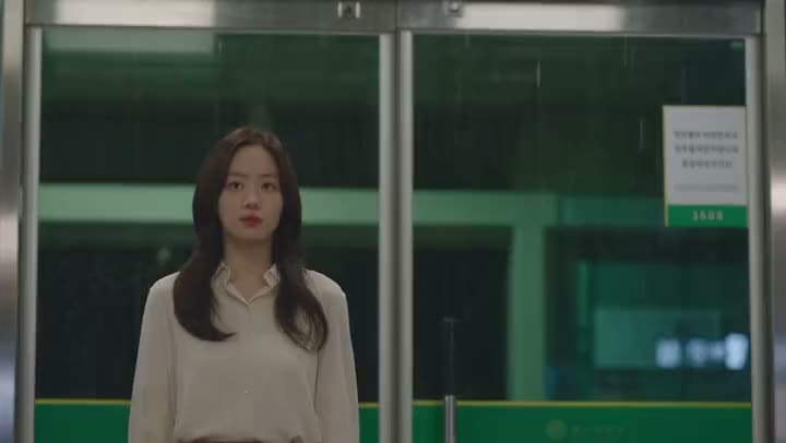 An AI video of a young woman in a cream blouse standing at the glass doors of a subway exit as rain falls outside.