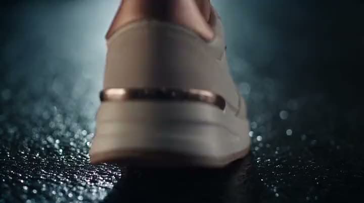 An AI sneaker ad close on the heel of a cream sneaker with a rose-gold stripe stepping onto a wet, glittering dark surface.