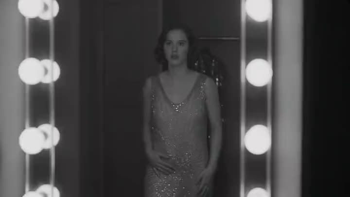 A black-and-white AI video of a woman in a beaded 1920s dress standing between rows of dressing-room mirror lights.
