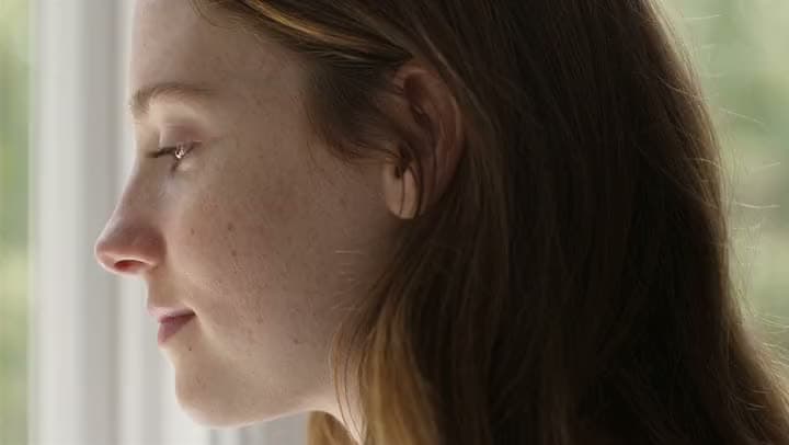 An AI video close-up of a young woman's freckled face in profile beside a bright window, with fine skin and hair detail.