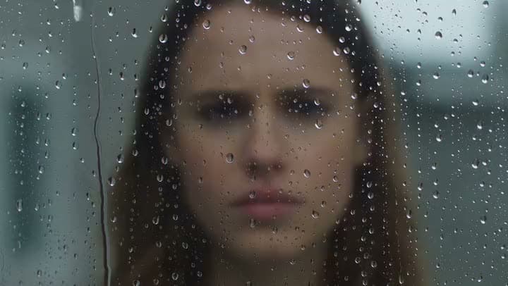 An AI video of a frowning young woman seen through a window pane covered in raindrops, her face softly out of focus.