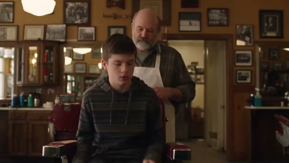 An AI video of a teenage boy in a striped hoodie sitting in a barber chair as an older bearded barber stands behind him.
