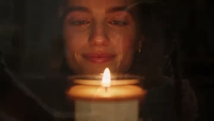 An AI candle commercial of a smiling woman with closed eyes leaning over a lit candle in a dark room, its flame glowing below her face.