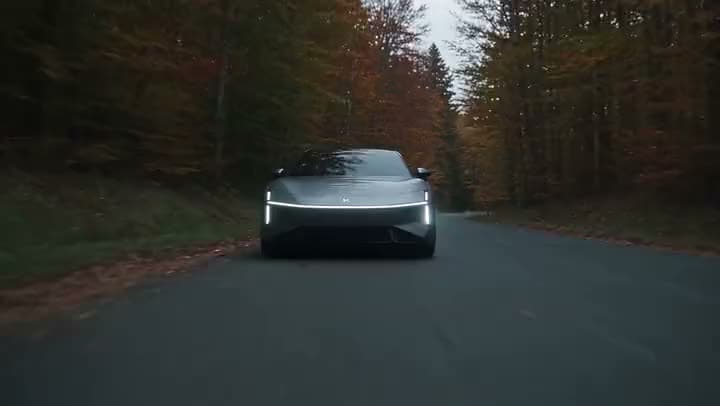 An AI car ad opening on a silver sports sedan driving toward the camera along a winding autumn forest road.