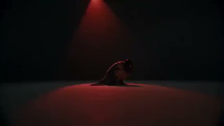 An AI video of a dancer crouched low on a bare stage, lit only by a single red spotlight from above.