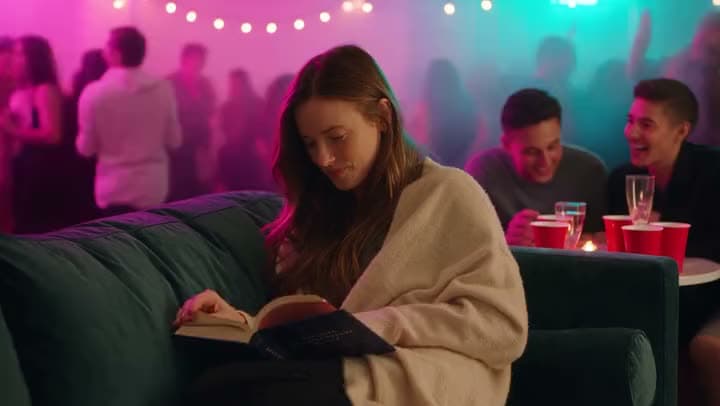 An AI video of a woman in a cream blanket reading a book on a sofa while a party with neon lights goes on behind her.