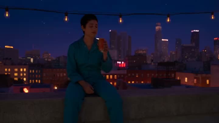 An AI video of a woman in a teal jumpsuit sipping a drink on a rooftop ledge under string lights and a night skyline.