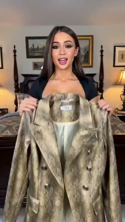 A vertical AI video of a woman in a bedroom holding up a snakeskin-print double-breasted blazer to the camera as she talks.
