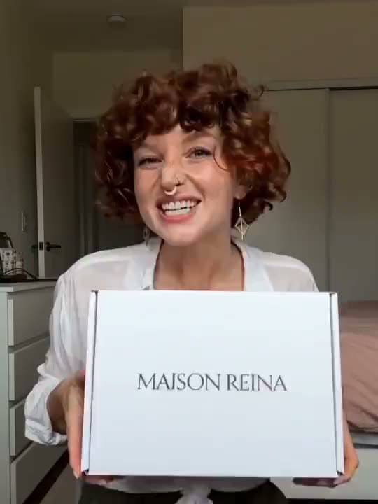 A vertical AI video of a smiling woman with curly auburn hair holding up a white MAISON REINA box in her bedroom.