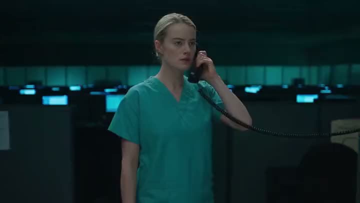 An AI video of a nurse in green scrubs holding a corded phone to her ear in a darkened hospital office full of monitors.