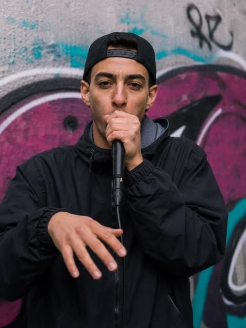 A portrait photo of a man in a backwards cap beatboxing into a handheld microphone against a graffiti-covered wall.