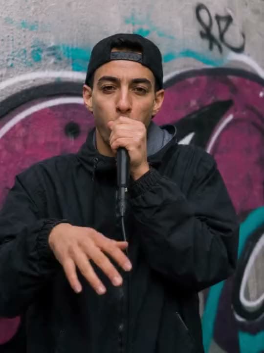 An AI video of a young man in a black cap and jacket beatboxing into a handheld microphone in front of a graffiti wall.