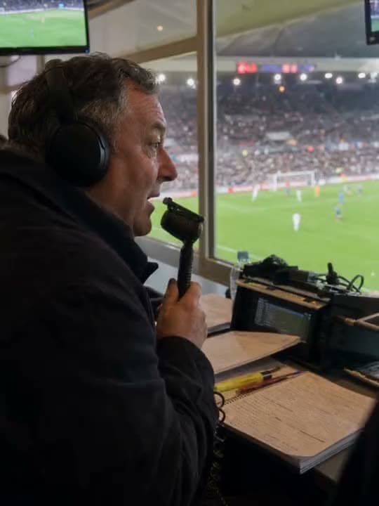 A vertical AI video of a sports commentator in headphones speaking into a handheld mic from a booth over a soccer match.