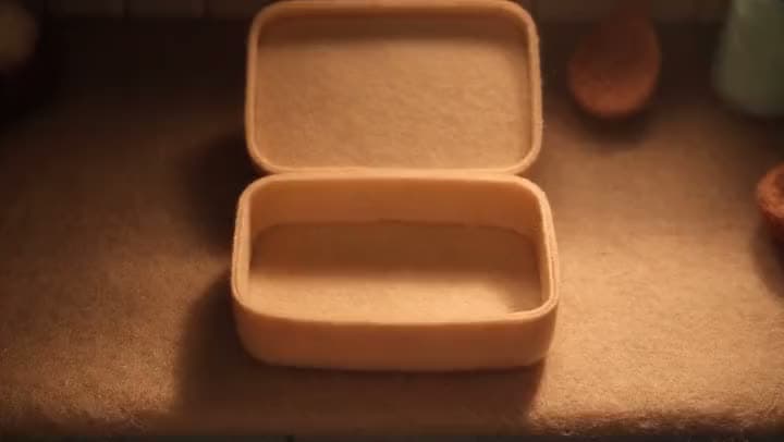 An AI stop-motion clip of an empty felt lunch box with its lid open, sitting on a felt table in warm, low light.